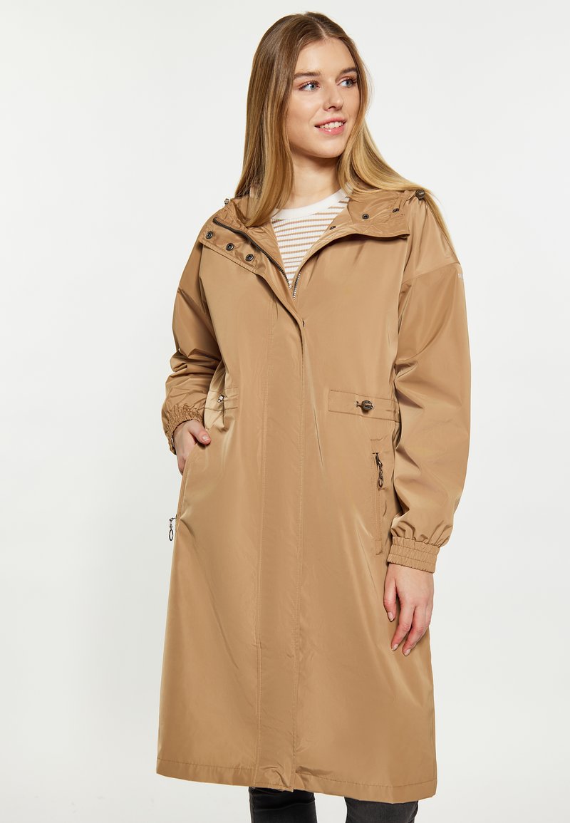 Beige vattentät kappa med huva, med dragkedjefickor, elastiska ärmslut och tryckknappsdetaljer, som sträcker sig till halva vaden. Slät textur.