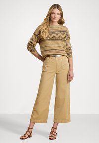 Beige patterned sweater with zigzag design, worn with light khaki wide-leg pants and brown strappy sandals. Model poses with hands on hips.