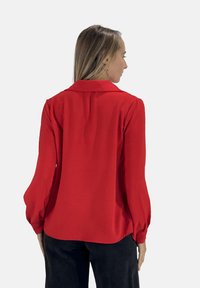 Blusa rossa con colletto, maniche lunghe e svasate, tessuto morbido e testurizzato, e orlo arrotondato, abbinata a pantaloni scuri.