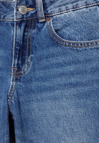 Kék farmernadrág közeli kép, amely az első zsebet, a fém gombot és a narancssárga varrás részleteit mutatja.