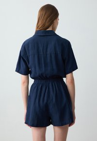 Jumper court bleu marine avec manches courtes, taille élastique et poches latérales. Fabriqué en tissu léger et texturé.