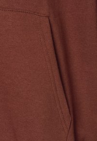 Primo piano di una tasca di una felpa con cappuccio marrone, con cuciture visibili e texture a maglia.