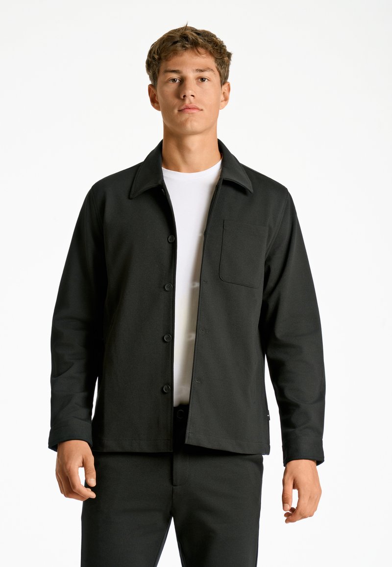 Chaqueta ligera negra de botones con cuello, bolsillo en el pecho y textura suave, combinada con pantalones a juego.