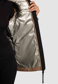 Schwarze Außenjacke mit metallisch goldener Fütterung. Verfügt über eine glatte Textur, kontrastierende sandfarbene Akzente und einen sichtbaren Reißverschluss mit Markenlogo.