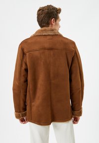 Mand iført en brun shearlingjakke med pelskrave og manchetter, stående med ryggen til og iført hvide bukser mod en hvid baggrund.