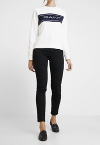 Witte sweater met donkerblauw GANT-logo over de borst, gecombineerd met zwarte skinny jeans en zwarte loafers. Gladde stof, ruime pasvorm.