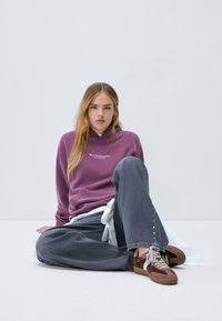 Jeune femme assise sur le sol, portant un sweat à capuche violet, un pantalon noir ample avec des clous et des baskets bordeaux avec des lacets clairs, sur un fond uni.