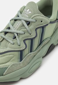 Zelené tenisky se semišovými a síťovinovými materiály, texturovanými detaily, třemi tmavě zelenými pruhy a stříbrnými logovými akcenty.