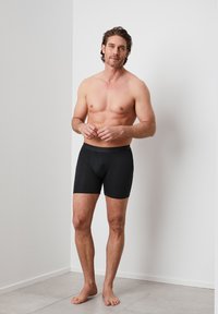 Schwarze Boxer-Briefs, die an einem Mann präsentiert werden, mit einem figurbetonten Design, glattem Stoff und einem mittelhohen Bund. Der Hintergrund ist minimalistisch und neutral.
