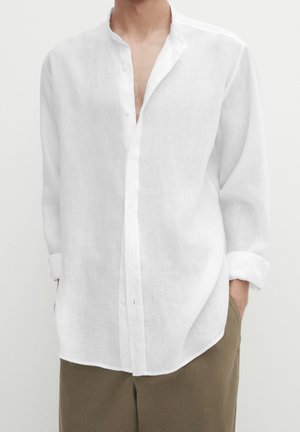 Homme portant une chemise en lin blanche ample à boutons avec les manches retroussées, associée à un pantalon marron, les mains dans les poches.