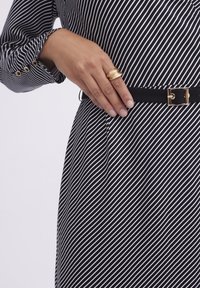 Czarna i biała sukienka w paski z długimi rękawami, wykończona czarnym paskiem z złotą klamrą i ręką ozdobioną złotym pierścionkiem.