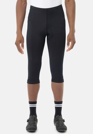 Schwarze, gekürzte Radlerhose, getragen von einer Person mit schwarzen Socken und schwarzen Radschuhen, stehend vor weißem Hintergrund.