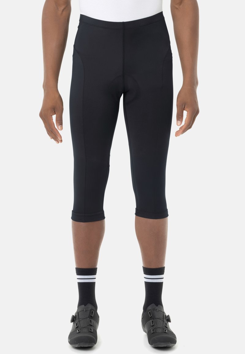 Pantaloncini da ciclismo neri tagliati indossati da una persona con calzini neri e scarpe da ciclismo nere, in piedi su uno sfondo bianco.