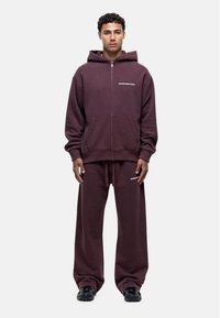 Haut à fermeture éclair bordeaux avec une poche kangourou, associé à un pantalon ample assorti. Présente un détail logo blanc sur la poitrine et le pantalon.