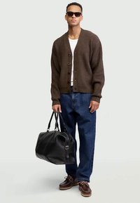 Uomo con occhiali da sole, cardigan marrone, camicia bianca, jeans blu, scarpe marroni, che tiene una borsa a tracolla in pelle nera davanti a uno sfondo semplice.