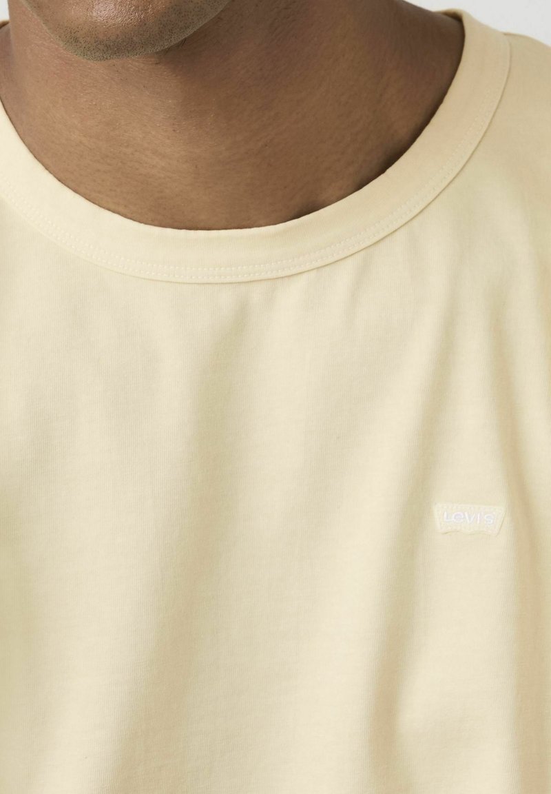 Primo piano di una persona che indossa una t-shirt Levi's tinta unita color crema, con scollo rotondo e piccolo logo sul petto.
