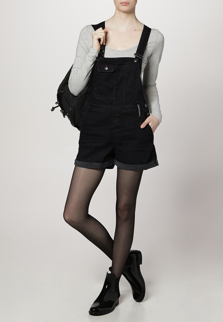 Personne portant une combinaison courte en denim noir, un t-shirt à manches longues gris, des collants noirs transparents, des bottines noires et un sac à dos noir.