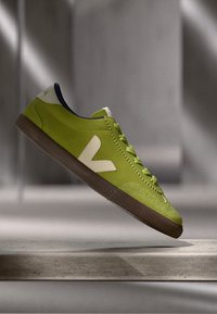 Roheline seemisneaker, millel on ümmargune varvas, valge V-logo, pruun kummitald ja tumesinine vooder üleval, tekstureeritud paeltega.