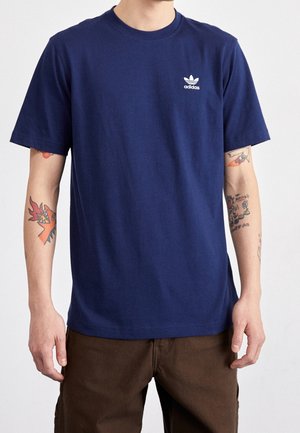 Mand iført en marineblå Adidas T-shirt med logo på brystet, brune bukser og synlige farverige tatoveringer på begge underarme.