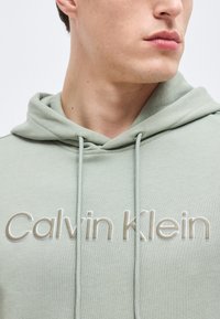 Jeune personne portant un sweat à capuche Calvin Klein vert clair avec cordons, cou visible et partie inférieure du visage tournée vers la droite.