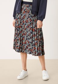 Jupe midi plissée à motif floral présentant des nuances de rouge, vert et marine, associée à des baskets blanches et un manteau bleu foncé.