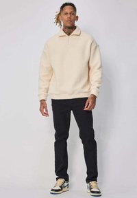 Pull en polaire couleur crème avec un col zippé à moitié, associé à un pantalon noir et des baskets beiges avec une touche de bleu.