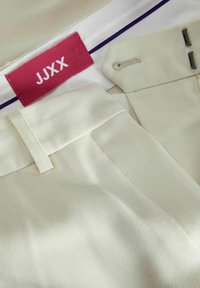 Beiga tygbyxor med en slät yta, rak framdel och en midjebandsetikett i röd med texten "JJXX" i vit.