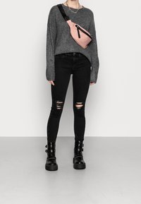 Grauer Strickpullover, schwarze ausgefranste Skinny Jeans und schwarze klobige Ankle Boots. Eine rosa Hüfttasche mit einem schwarzen Riemen wird diagonal über den Körper getragen.