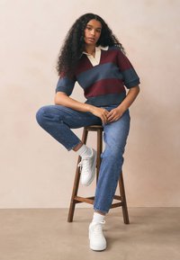 Megztinis su bordo, mėlynomis ir kreminėmis juostelėmis, trumpomis rankovėmis ir V formos iškirpte. Derinamas su mėlynomis džinsais ir baltais sportbačiais, sėdint ant kėdutės.