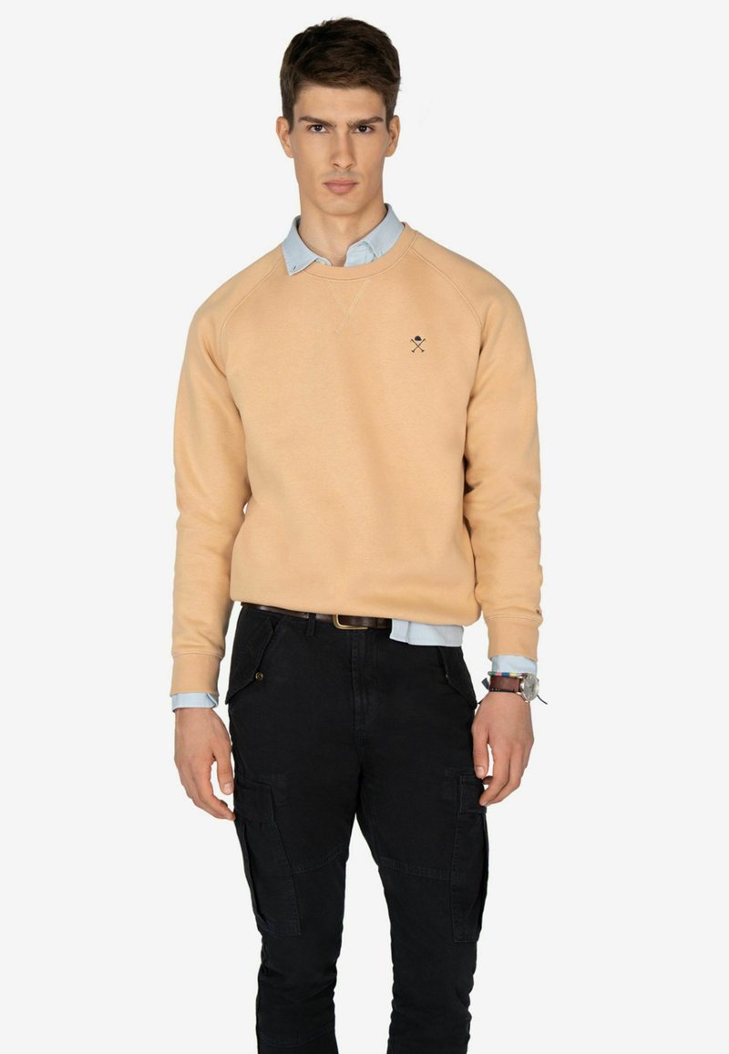 Licht perzik kleurige sweatshirt met raglanmouwen, ronde halslijn en klein zwart logo op de borst. Draag het over een lichtblauw overhemd met kraag en zwarte broek.