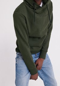 Felpa verde oliva con collo alto, lacci e vestibilità comoda, abbinata a jeans azzurri chiari. Tessuto morbido con un design minimalista.