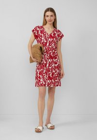 Rotes kurzes Kleid mit einem weißen Blattmuster, V-Ausschnitt und kurzen Ärmeln, tailliert in der Taille. Das Model hält eine beige geflochtene Tasche und trägt weiße Sandalen.