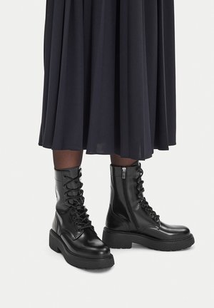 Czarne skórzane botki na sznurowadła z bocznym zamkiem, noszone z czarnymi przezroczystymi rajstopami i długą czarną spódnicą na jasnym tle.