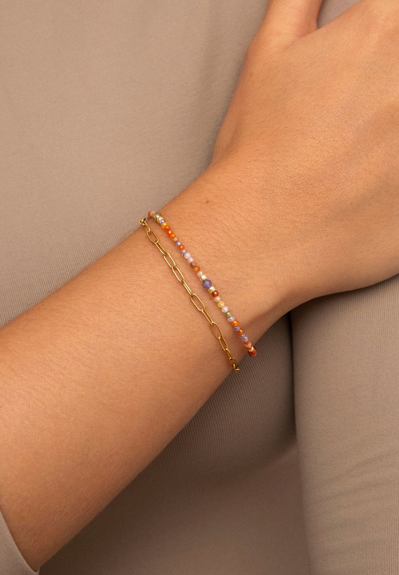 Bracciale in oro abbinato a un bracciale di perline colorate, con perline arancioni, viola e pastello su uno sfondo di tonalità della pelle neutra.