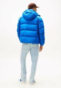 Blauwe pufferjas met een capuchon, gevoerde textuur en elastische manchetten. Gecombineerd met lichtblauwe jeans en witte sneakers. Bruine pet op het hoofd.