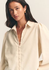 Cream-colored blouse with a buttoned front, collar, and puffed sleeves. Soft fabric texture with rounded buttons detailing.