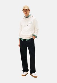 Joven vestido con una gorra beige, sudadera blanca, camiseta a rayas verdes, pantalones negros y zapatillas blancas, de pie con las manos en los bolsillos.
