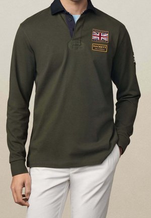 Homme portant un polo à manches longues vert foncé avec un col noir, écusson du drapeau britannique, et un pantalon blanc, debout avec une main dans la poche.