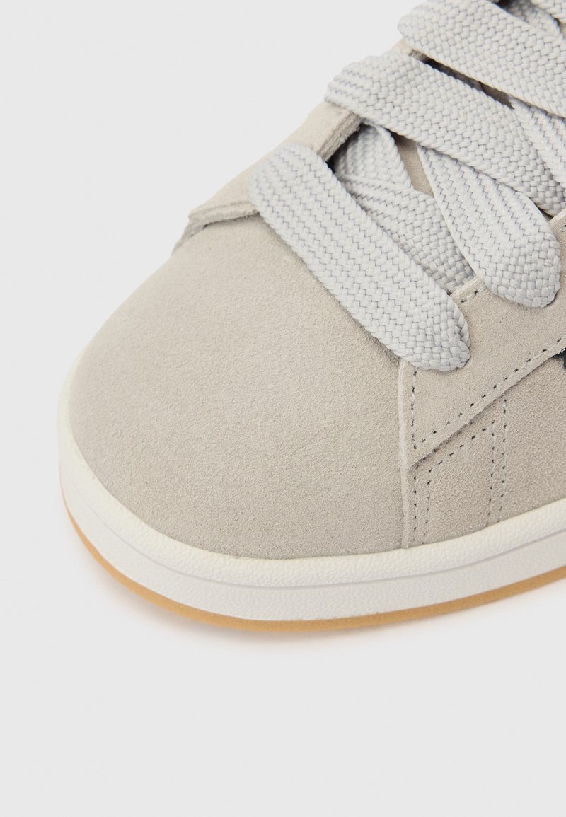 Grå mockasinsneaker med rund tå, texturerade grå snören och vit gummisula. Har minimalistisk design med subtila söm-detaljer.