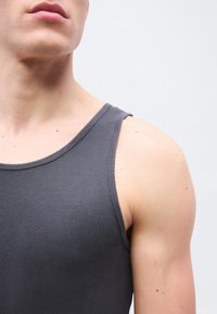 Κοντινό πλάνο του άνω μέρους του κορμού ενός ατόμου που φοράει γκρι σκούρο ριμπαριστό αμάνικο τοπ σε απλό φωτεινό φόντο.