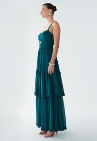 Abito lungo turchese con corpetto aderente, spalline regolabili e orlo a balze a più livelli. Realizzato in tessuto leggero con una consistenza morbida.