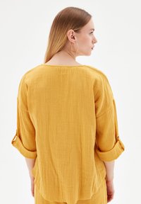Mosterdgele blouse van textuurstof, met opgerolde mouwen met knopen en een losse pasvorm, met een rechte rugontwerp.
