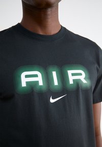 Juodas medvilninis marškinėliai su žaliai balta "AIR" grafika, turinčia banguotas linijas ir mažą baltą Nike swoosh logotipą.