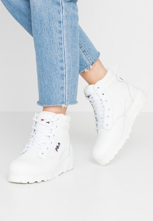 Gambe con jeans corti azzurri e sneakers bianche alte FILA con suole spesse su uno sfondo chiaro.