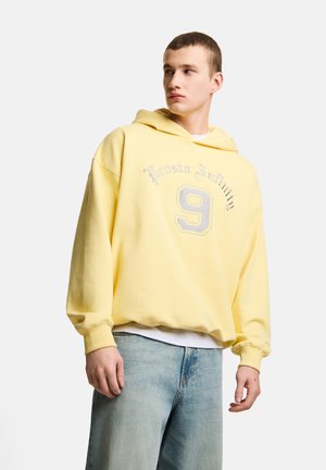 Młody mężczyzna ubrany w luźną żółtą bluzę z kapturem z napisem „Prosto Infinity 9”, jasnoniebieskie jeansy, stojący na jednolitym białym tle.