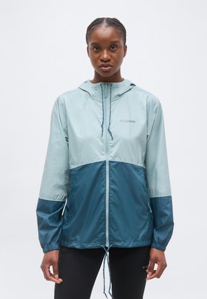 Junge Frau, die nach vorne blickt und eine zweifarbige Columbia-Windjacke in Hellblau und Türkis mit Kapuze, Frontreißverschluss und Kordelzug trägt.