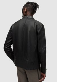 Schwarze Lederjacke mit taillierter Passform, minimalen Nähten und Reißverschlussmanschetten. Verfügt über einen Stehkragen und eine glatte Textur.