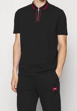 Mann trägt schwarzen Kurzarm-Poloshirt mit rotem Besatz und Reißverschluss, kombiniert mit schwarzen Hosen mit rotem und weißem Hugo-Logo.