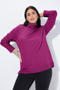 Donna con capelli lunghi e scuri che indossa un maglione dolcevita magenta e pantaloni neri, posando con una mano sui fianchi e l'altra dietro la testa.