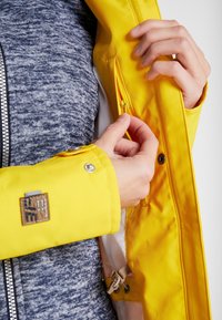 Osoba zsuwa wewnętrzną kieszeń jasnotyellow kurtki, nosząc pod nią niebieską strukturę zapinaną na zamek.